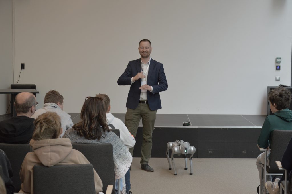 Na zdjęciu widać mężczyznę w granatowej marynarce. Gestykuluje, prawdopodobnie prowadzi wykład. Obok niego stoi niewielki metalowy robot w kształcie psa bez głowy. Przed mężczyzną widać siedzącą grupę słuchaczy. Zdjęcie: MBP.