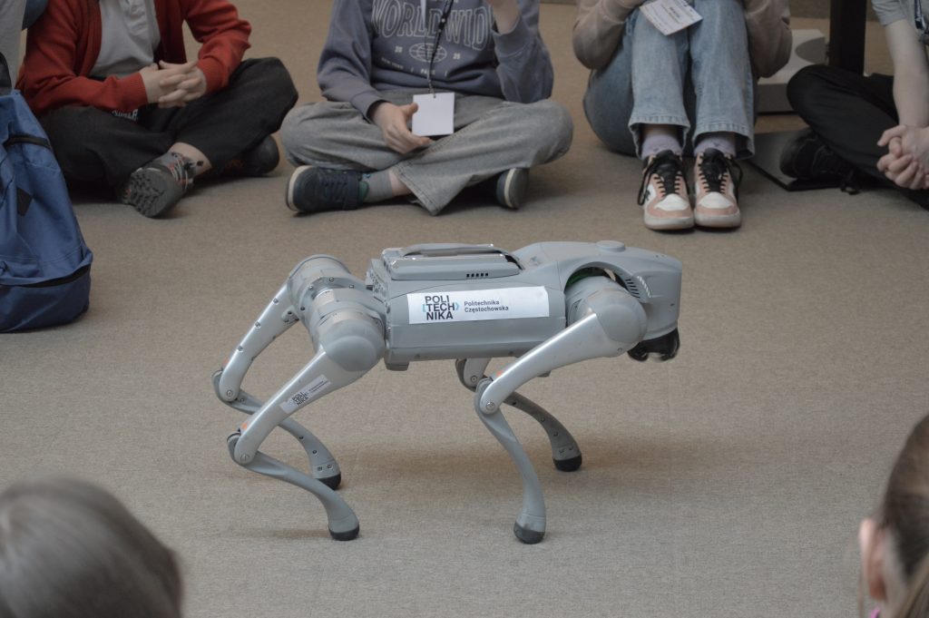 Zdjęcie przedstawiające metalowego robota w kształcie psa. Jego nogi są wygięte w łuk w stronę tyłu psa. Na boku robota pisać logo oraz nazwę Politechniki Częstochowskiej. W tle widać nogi dzieci siedzących na podłodze. Zdjęcie: MBP.