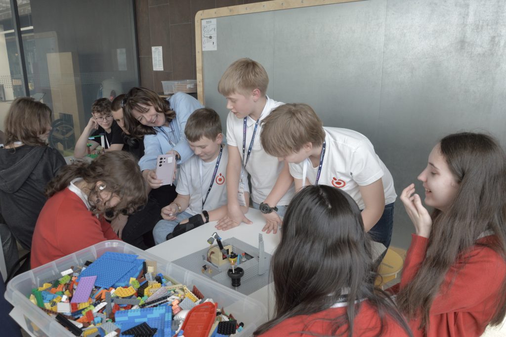 Dwa stoły z klockami LEGO, przy których siedzą dwie grupy nastolatków. W tle metalowa tablica. Zdjęcie: MBP.